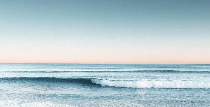 Escena serena del océano con olas suaves que se dirigen hacia la orilla. El cielo está despejado con un degradado de azul claro en la parte superior a un suave color naranjo cerca del horizonte, sugiriendo el amanecer o el atardecer