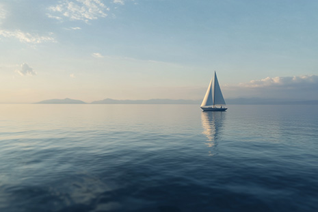 Velero con velas blancas navegando en aguas tranquilas y reflectantes bajo un cielo despejado. El horizonte es visible en el fondo con montañas distantes.